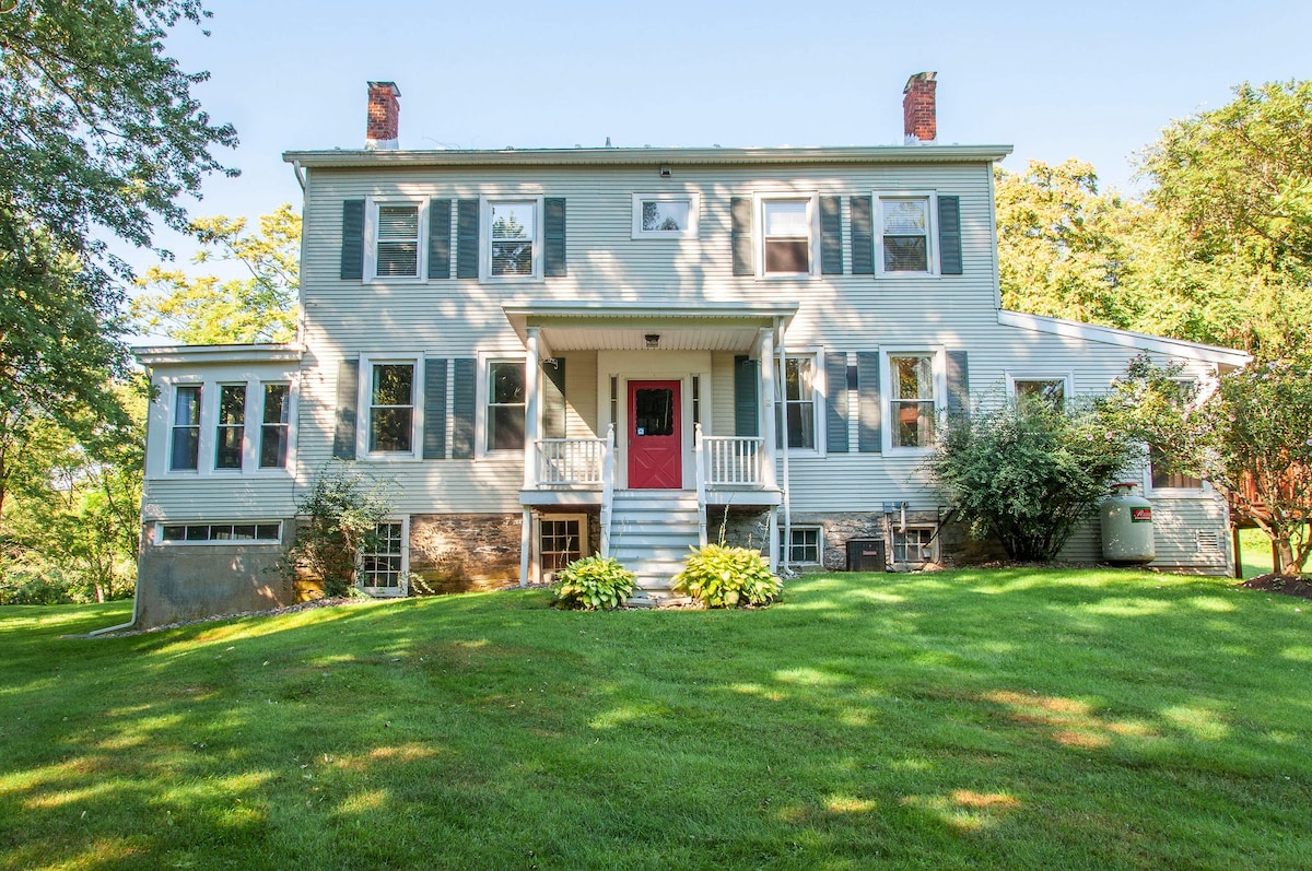 The exterior of the historic family home is shown, featuring a light grey facade with blue shutters and a red front door. The welcoming entrance includes steps and a porch, surrounded by lush green grass and trees, set on a gently sloping lawn.