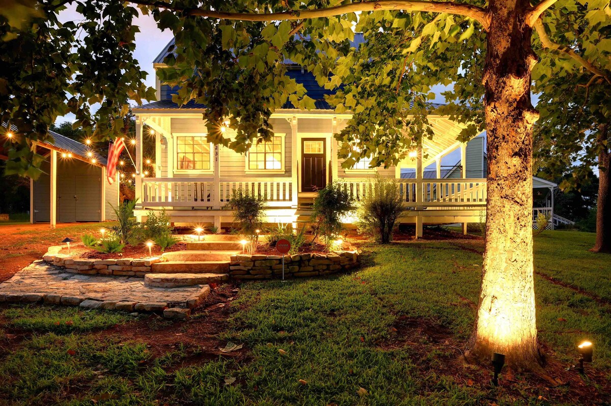The exterior of a charming farmhouse is illuminated by soft outdoor lighting, creating a warm ambiance. A spacious wrap-around porch and stone steps lead to the entrance. Colorful shrubs and a large tree add to the inviting landscape, providing a serene outdoor space.