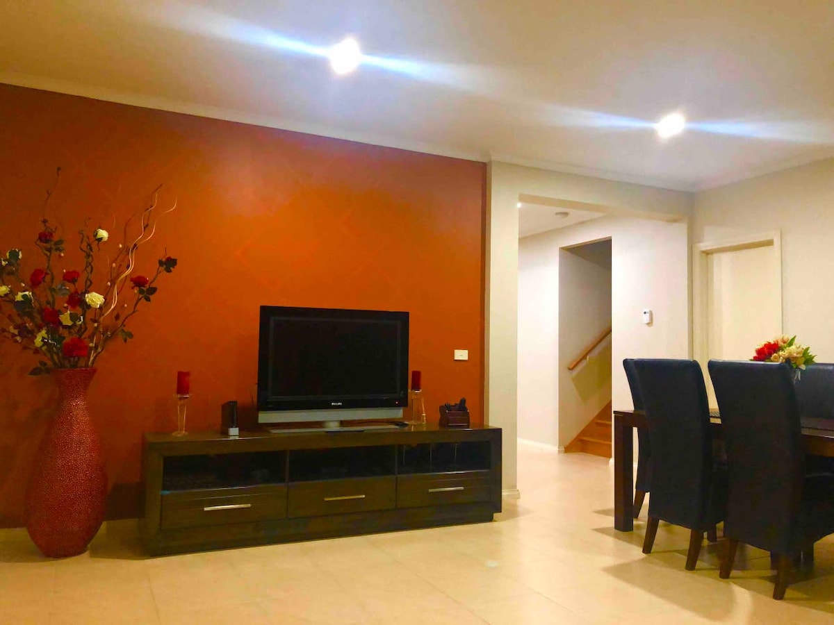 A cozy living area features a modern television mounted on a dark wood console, complemented by decorative elements including a tall vase with an assortment of flowers. Soft lighting enhances the warm orange accent wall, while a dining area with dark chairs is visible in the background.