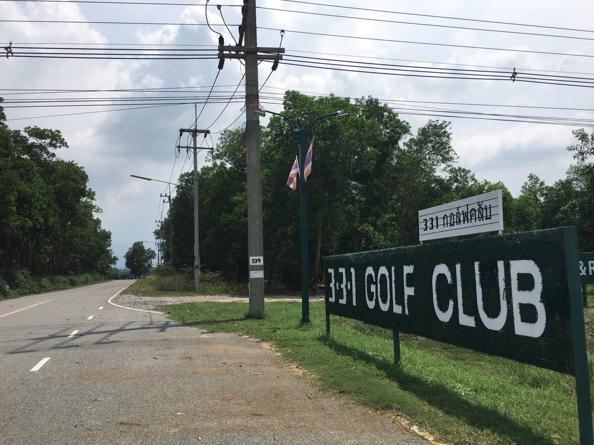 A sign for 331 Golf Club is prominently displayed beside a road, with greenery in the background. The area appears peaceful, with power lines overhead and a clear sky visible. The sign is painted in bold letters, making it easily legible from a distance.