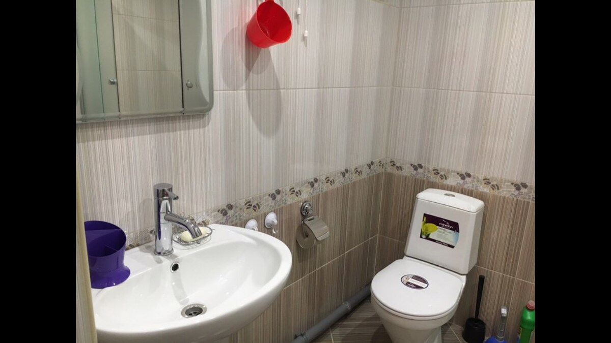 A minimalist bathroom features a wall-mounted mirror above a sink with a modern faucet. A toilet is positioned adjacent to the sink, while a tiled border accents the lower wall. Cleaning supplies are visibly organized near the floor.