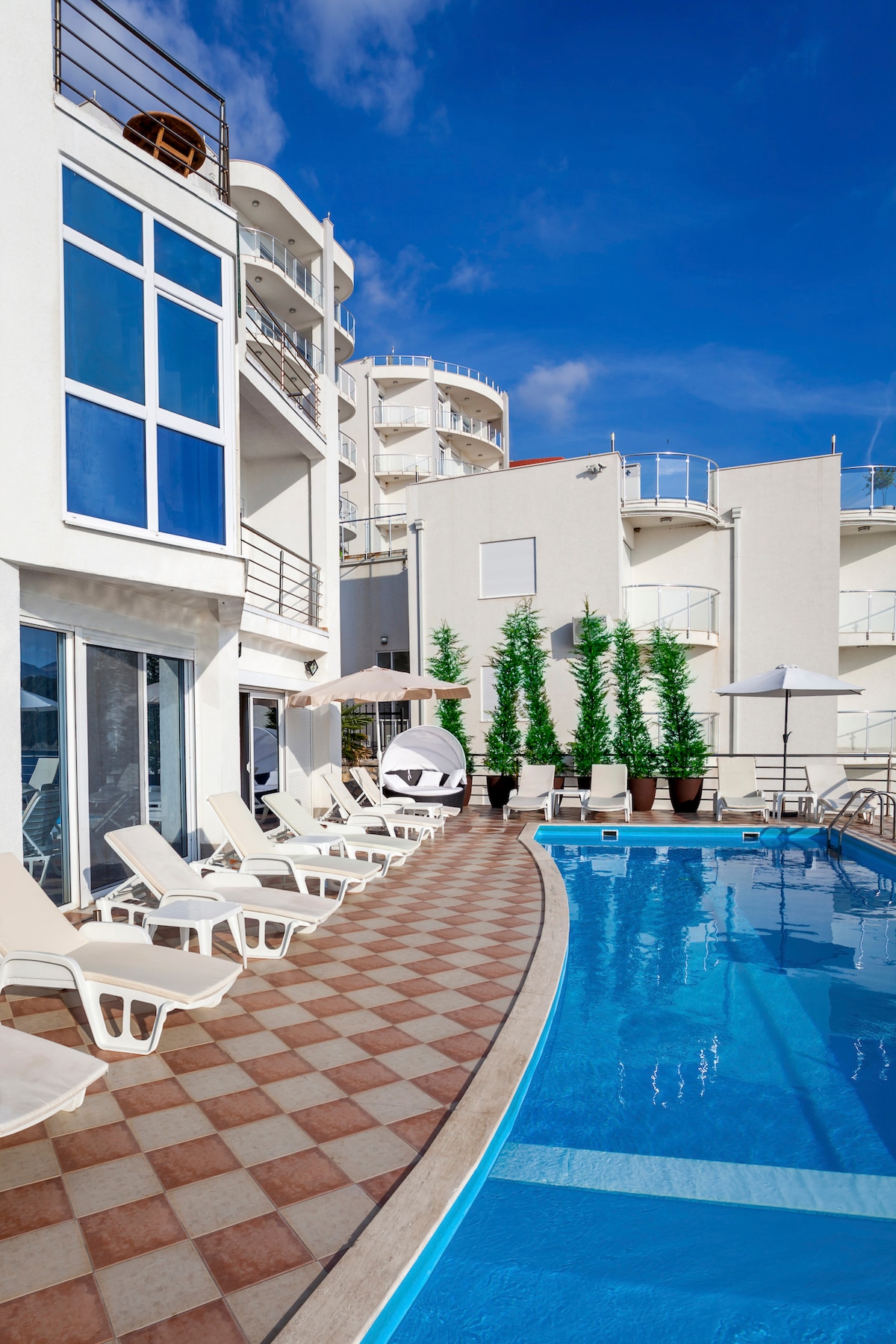 The outdoor area features a large swimming pool bordered by lounge chairs arranged along a patterned tiled deck. Towering green plants provide a touch of nature, while multi-story buildings with large glass windows reflect the blue sky.