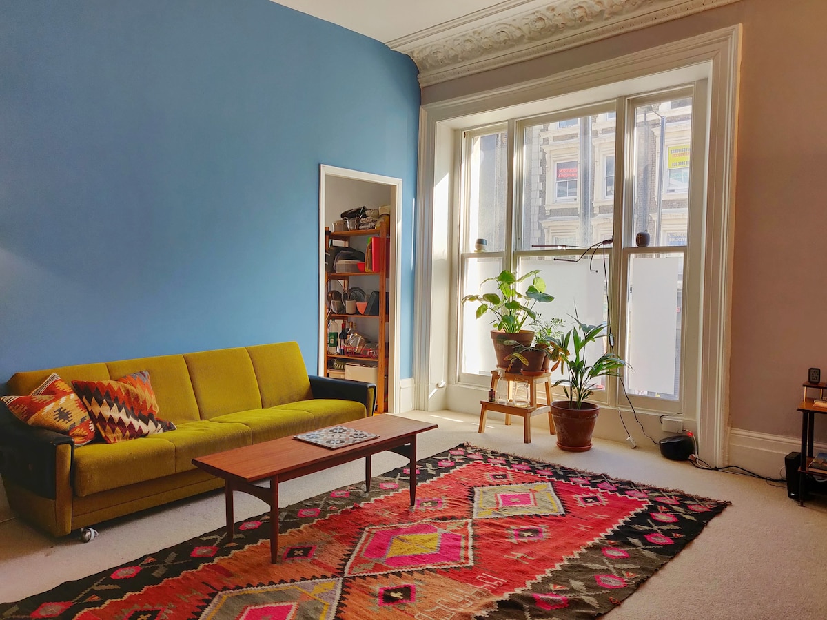 A spacious living area features a vibrant green sofa and a wooden coffee table on a colorful patterned rug. Large windows allow natural light to illuminate the room, highlighting a potted plant positioned nearby and a bookcase filled with items against the blue accent wall.