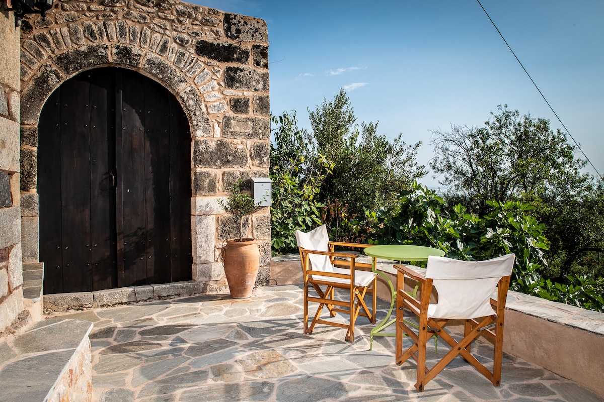 An outdoor terrace features a stone-paved floor and a large wooden door built into the ancient stone wall. Two wooden chairs with white fabric cushions are positioned around a small green table, surrounded by lush greenery and distant olive trees.