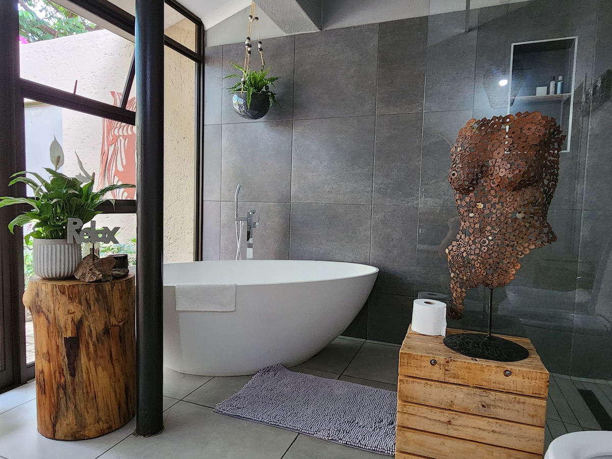 An exceptionally spacious bathroom is displayed, featuring a sleek, freestanding bathtub and a modern sculpture. Natural light filters through large glass windows. Decorative plants are positioned on a wooden stump, alongside a neatly arranged roll of toilet paper on a wooden crate.