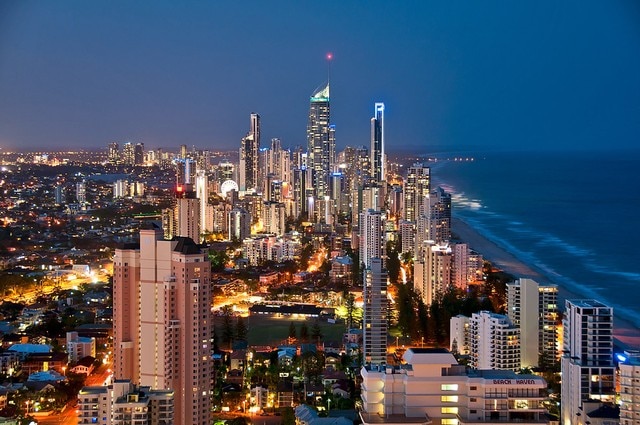 The city skyline of Broadbeach is illuminated at night, showcasing a mix of high-rise buildings and coastline. The ocean is visible to the right, with the lights reflecting off the water. The vibrant city atmosphere is highlighted by glowing structures along the shore.