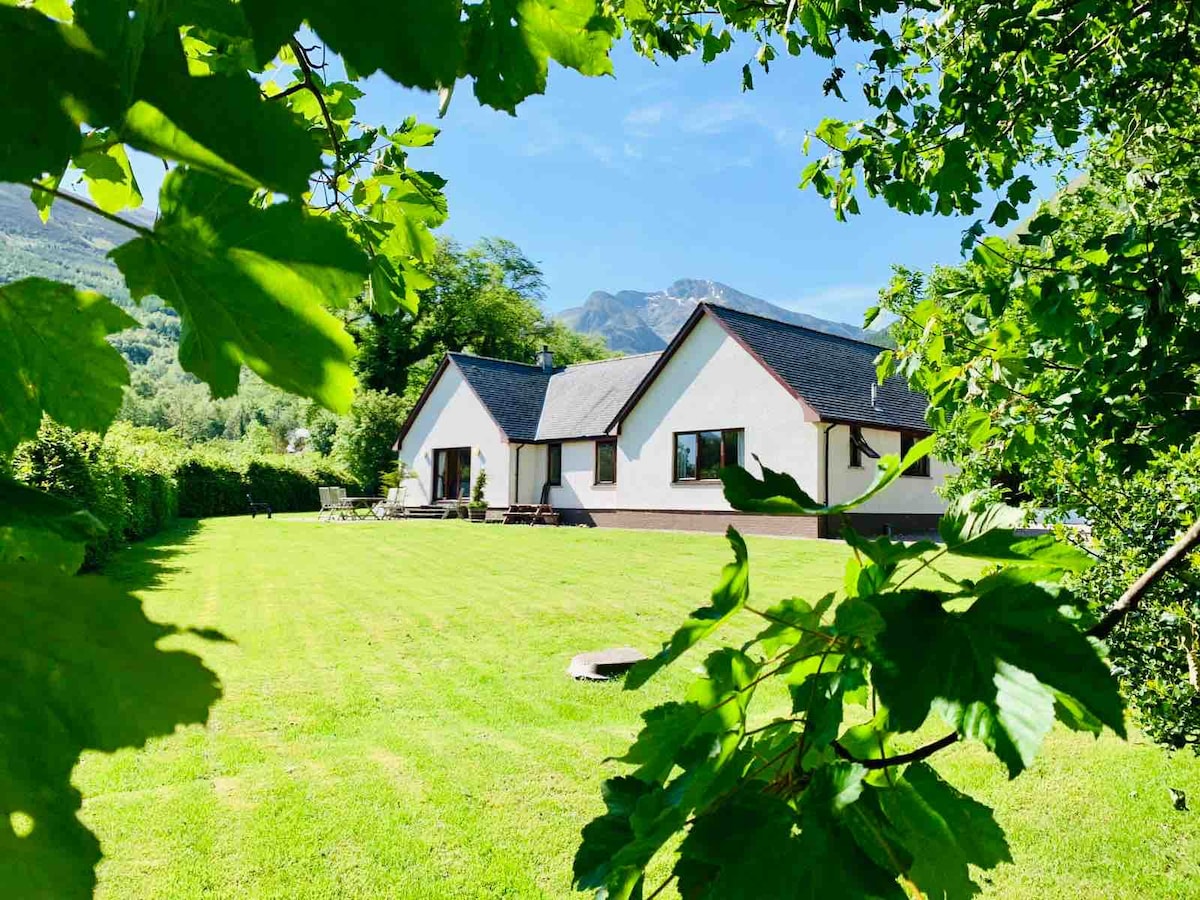 The exterior of a spacious lodge is surrounded by lush greenery. Large windows face a well-manicured lawn, while the sloping hills in the background suggest a serene mountainous landscape. The structure features a combination of white and dark roofing, providing a harmonious appearance.