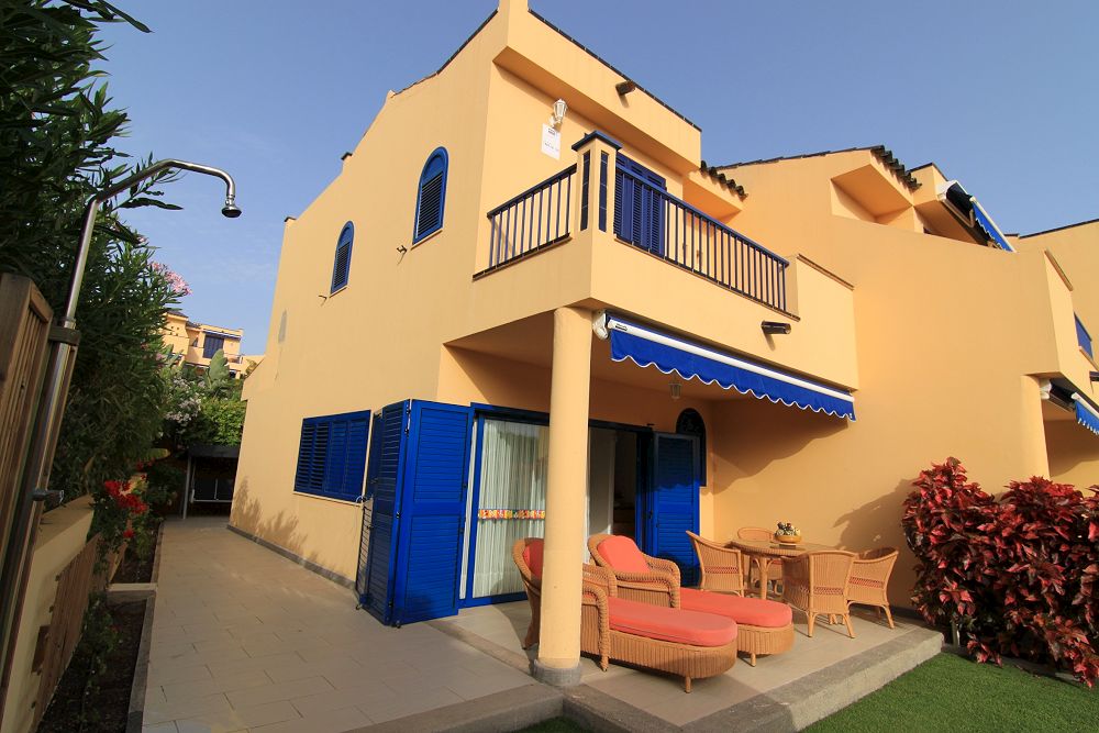 The exterior of a two-story duplex features yellow walls and blue shutters, creating a vibrant look. A broad patio area is shown, furnished with a table and chairs, alongside a comfortable seating arrangement. Lush greenery surrounds the space, enhancing the outdoor ambiance.