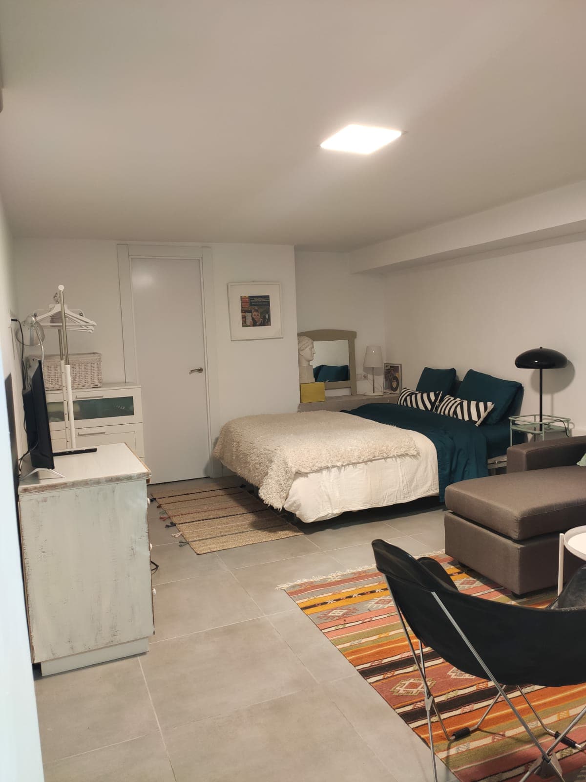 A modern studio space is presented with a spacious bed dressed in light linens. Comfortable seating areas include a plush armchair and a stylish sofa. A sleek television and decorative art pieces add to the cozy ambiance, with soft lighting provided by a standing lamp.