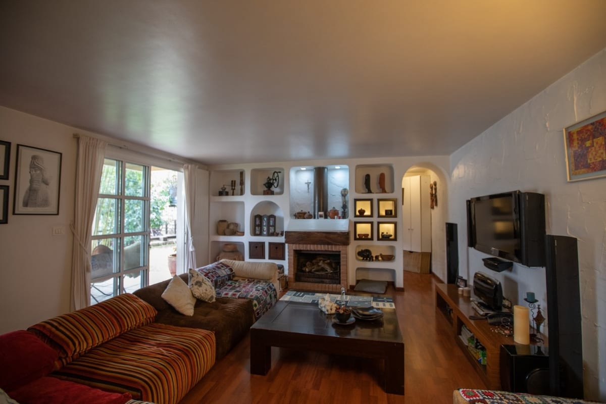 A welcoming living room features comfortable sofas with varied patterns and a central coffee table. A television is positioned on one wall, while a fireplace adds a cozy touch. Light enters through large glass doors, leading to the outdoor space, enhancing the room's brightness.