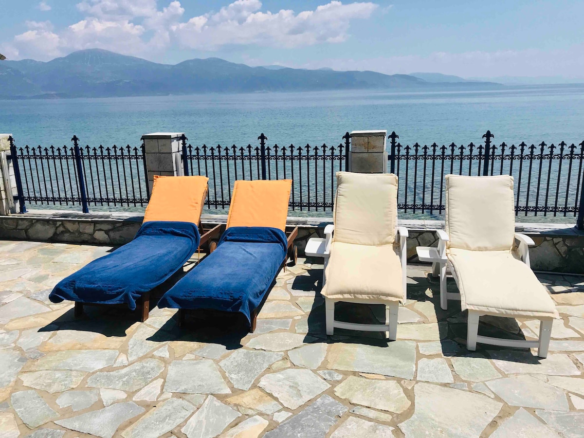 Four sun loungers are positioned on a stone terrace overlooking the sea. Two loungers are covered with blue and orange towels, while the other two are adorned with cream-colored fabric. A decorative metal fence frames the view of the calm waters and distant mountains.