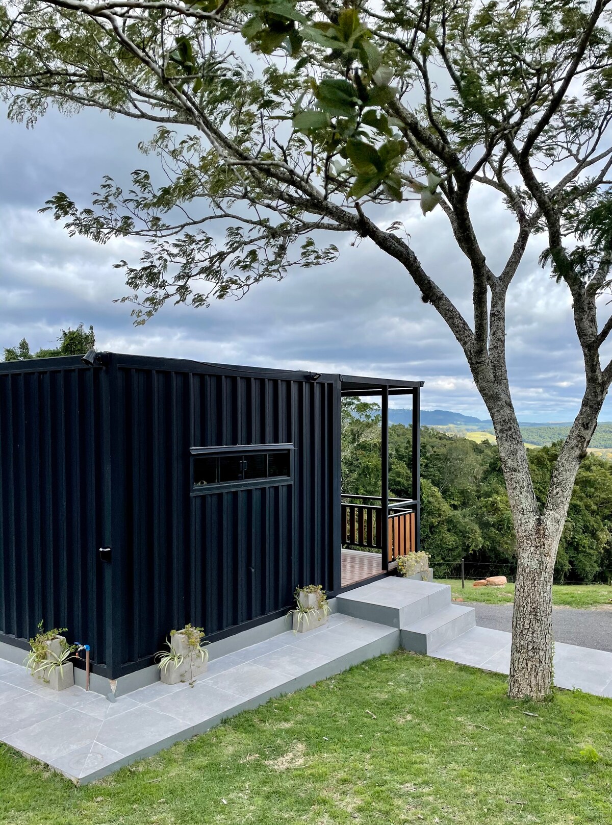 A modern container house is showcased, nestled among greenery. The dark exterior contrasts with the surrounding natural landscape. A set of stairs leads to a small deck, where views of the mountains and trees can be enjoyed. Potted plants add a touch of life to the setting.