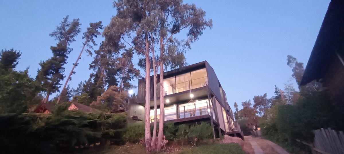A two-story house is set among tall eucalyptus trees at dusk. The structure features large glass windows that reflect the evening light. A wooden deck extends outward, providing an inviting entrance. Surrounding greenery adds to the natural landscape, enhancing the serene atmosphere.