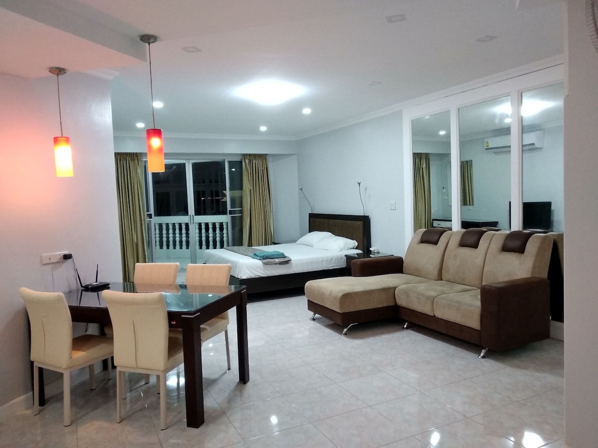 A spacious living area includes a comfortable sofa, a dining table with chairs, and a double bed visible through sliding glass doors. Natural light fills the room, accentuated by light-colored walls and elegant curtains, providing a cozy and inviting ambiance.