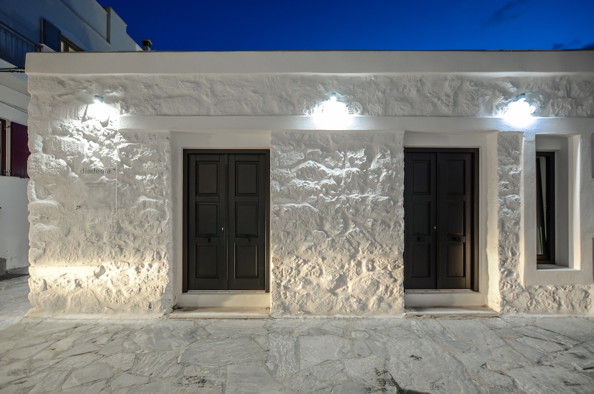 A newly constructed stone building is illuminated by soft outdoor lighting at dusk. The exterior features three dark wooden doors set against a textured white stone wall, complemented by a paved stone walkway in front.