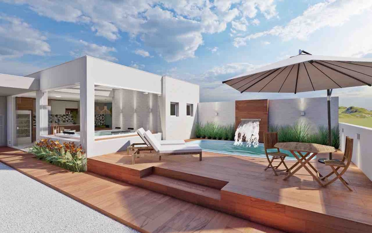 An outdoor area is presented with a sheltered dining space and a wooden deck. Lounge chairs are positioned near a tranquil pool featuring a small waterfall. An umbrella provides shade, while greenery and flowers enhance the environment, creating a relaxed and inviting setting.