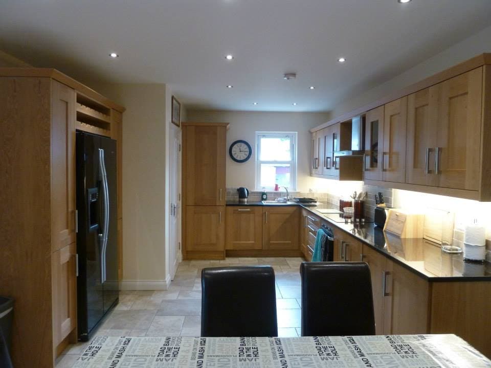 A modern kitchen displays wooden cabinetry complemented by dark countertops. Stainless steel appliances, including a refrigerator, are present. A dining table with four chairs is positioned in the foreground. Soft lighting enhances the spacious design, while a window allows natural light to fill the room.