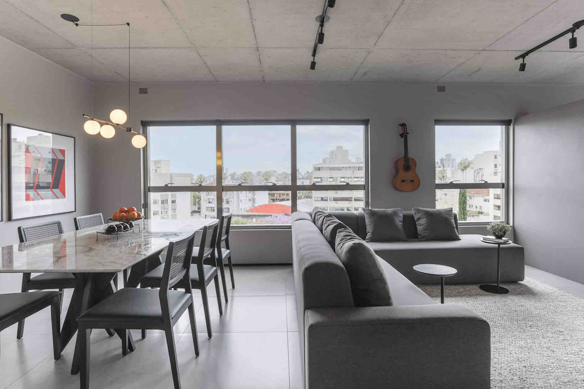 A modern living area is furnished with a gray sectional sofa and a small side table. A dining table with black chairs is positioned nearby. Large windows offer a view of the city skyline, allowing natural light to brighten the space.