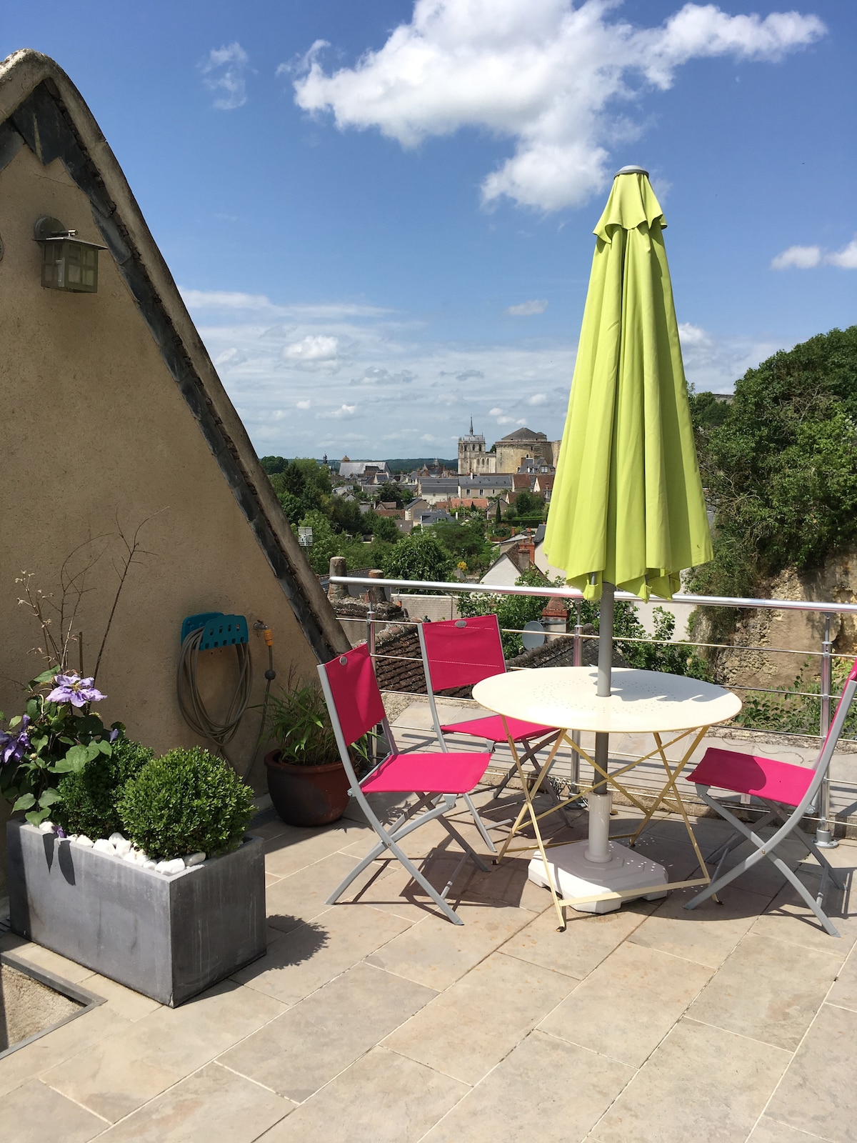 An outdoor terrace is presented, featuring a small round table surrounded by four vibrant chairs. A large green umbrella provides shade, while potted plants add greenery to the space. Scenic views of nearby buildings and trees stretch into the distance under a partly cloudy sky.