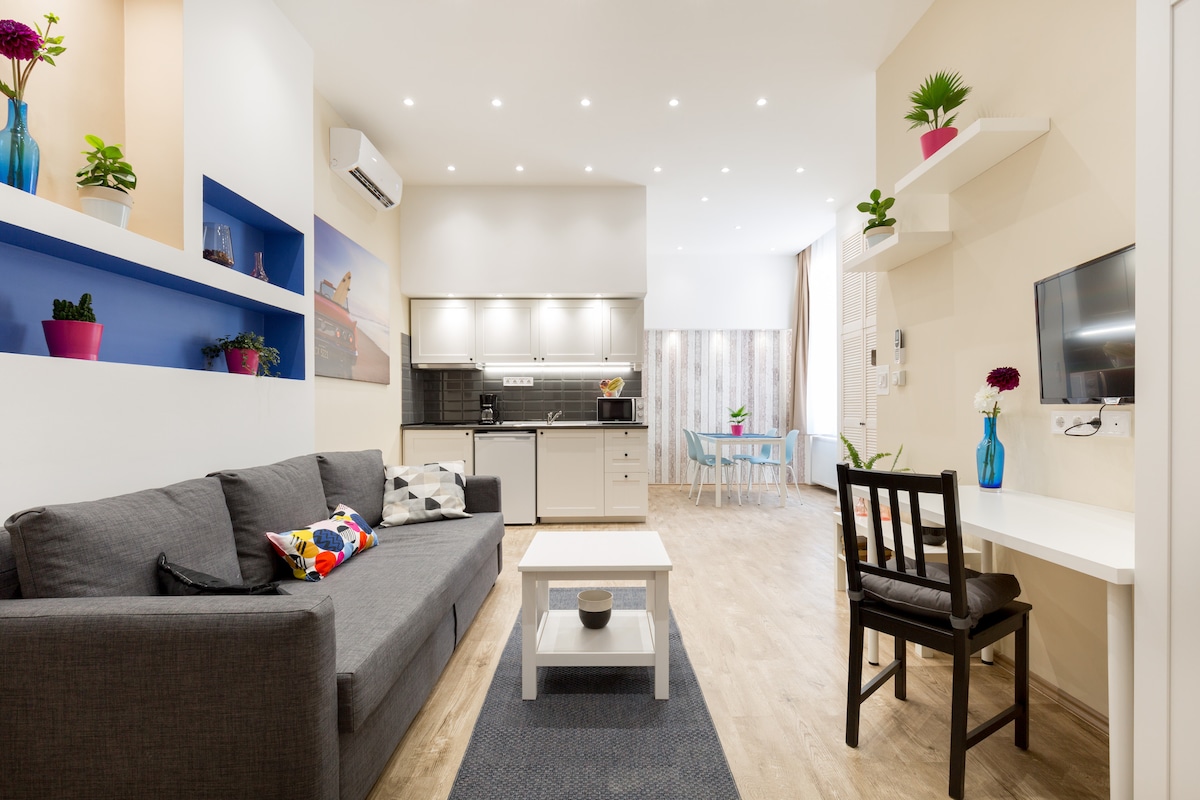 A modern studio living area is presented, featuring a gray sofa adorned with colorful cushions. Shelves with potted plants are visible, extending towards a well-equipped kitchen. A dining table with chairs is positioned near a window, while a flat-screen TV is mounted on the wall.