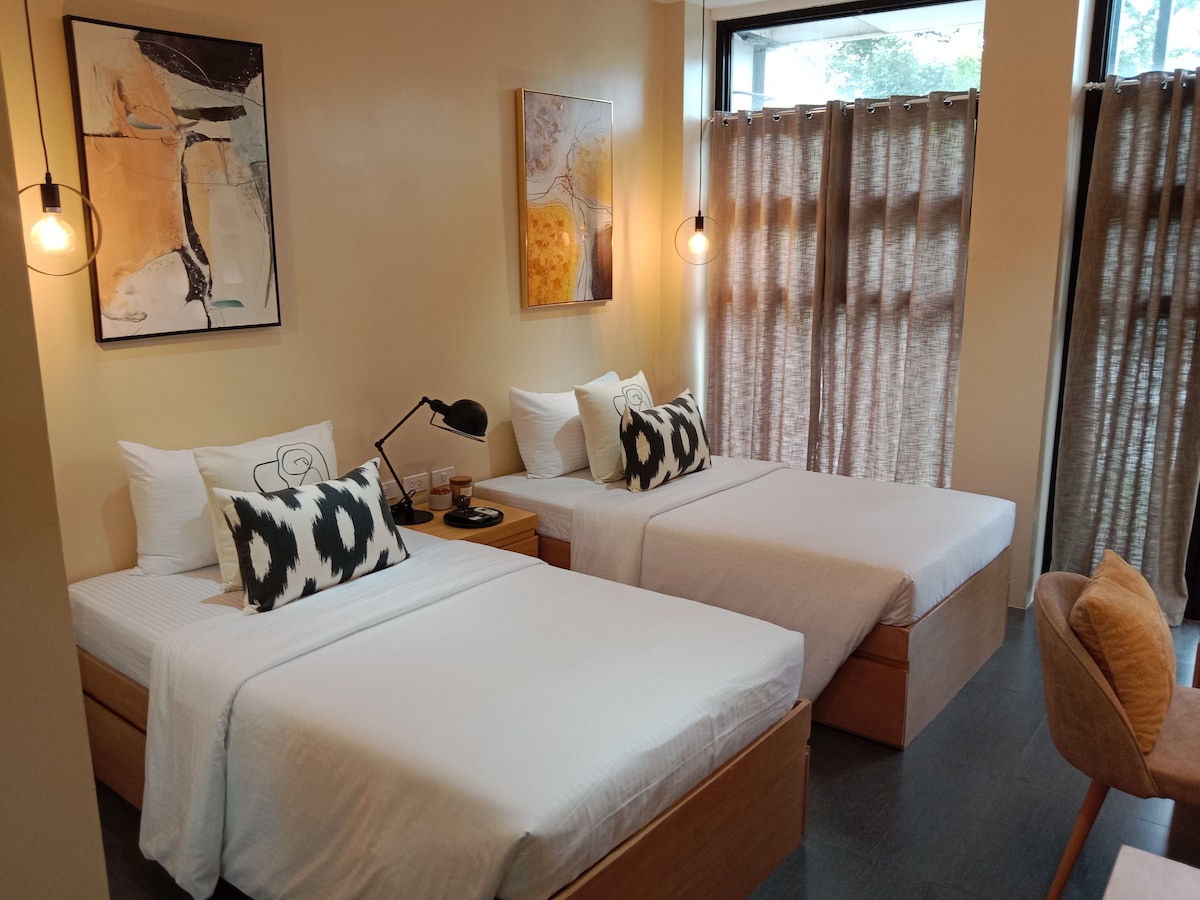 A well-lit guest room features two spacious beds with neutral bedding and decorative throw pillows. Natural light filters through sheer curtains, illuminating modern artwork on the walls. A sleek bedside lamp and a small table complement the minimalist design, creating a relaxing environment.