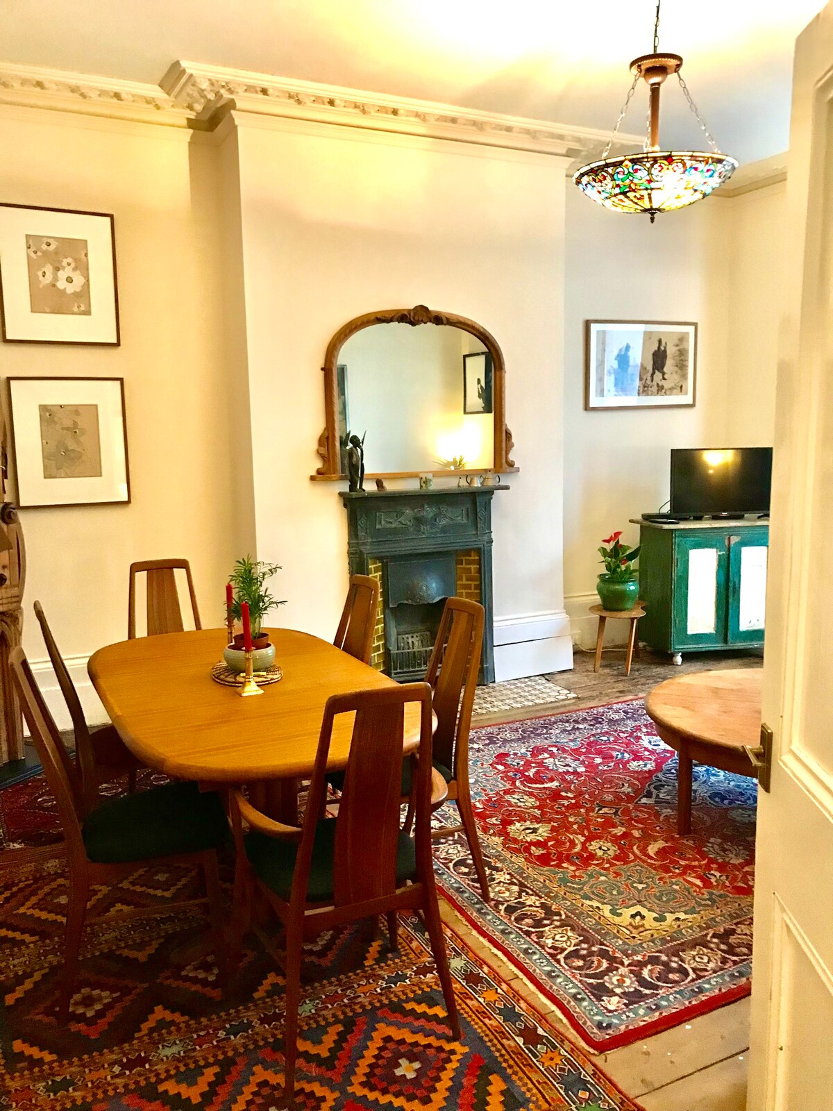 A dining area features a wooden table surrounded by six chairs, complemented by a colorful Persian rug. A vintage mirror and small fireplace are located against the wall, with framed artwork hanging nearby. A television stands on a green cabinet in the corner.