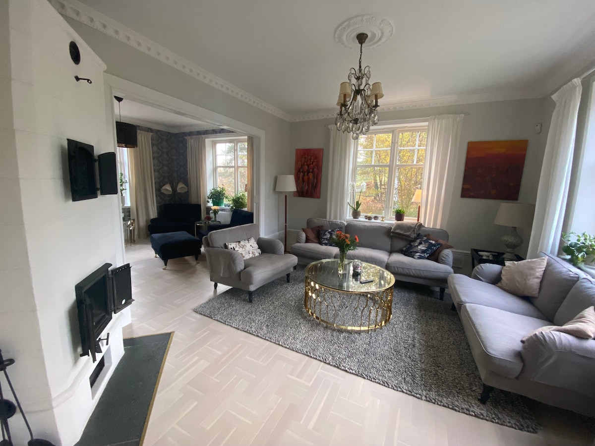 A spacious living area is highlighted by light grey sofas surrounding a circular gold coffee table, set atop a textured rug. Large windows allow natural light to fill the room, complemented by elegant lighting fixtures and tasteful artwork on the walls.