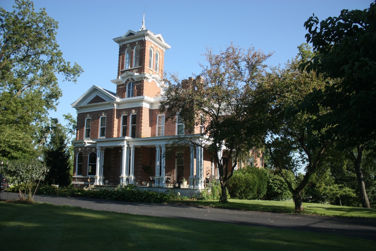 The grand Italianate villa is framed by lush greenery, showcasing its distinctive brick facade and intricate architectural details. A spacious porch is supported by tall columns, while trees provide natural shade, enhancing the overall charm of the property.