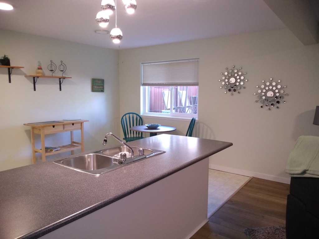 A bright kitchen area is featured with a spacious countertop and stainless steel sink, overlooking a small dining space. Two green chairs are paired with a circular table by a window. Decorative mirrors and shelves enhance the welcoming feel of the room.