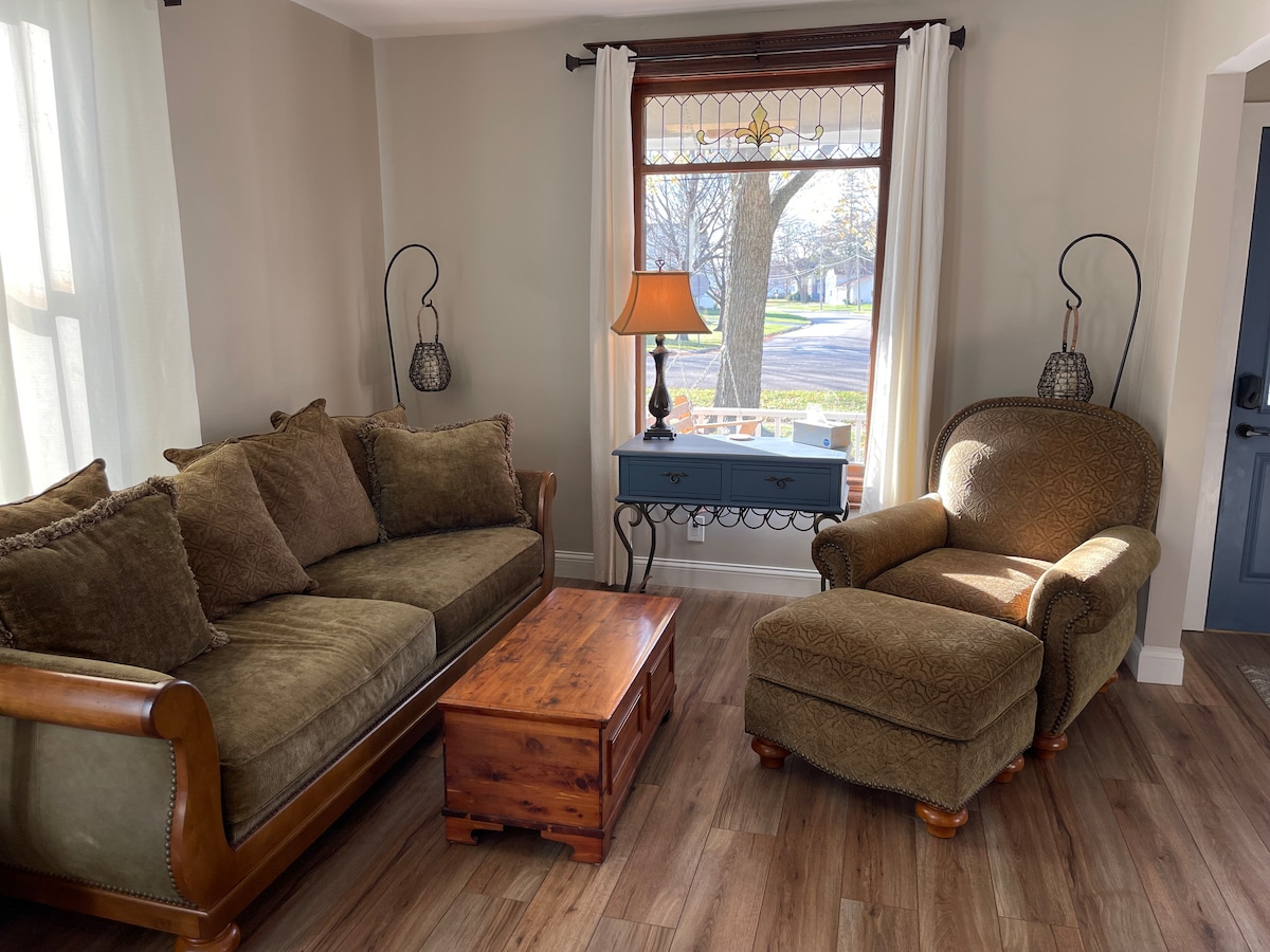 A cozy living area is presented with a plush brown sofa and an upholstered armchair, paired with a small wooden coffee table. Natural light enters through a large window, featuring decorative glass accents, while a lamp adds soft illumination to the space.