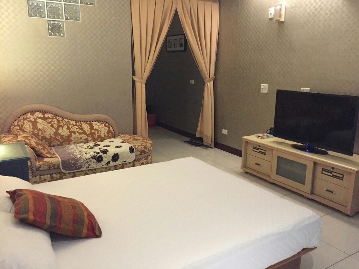 A comfortable room features a spacious bed with clean linens and a decorative pillow. A cozy seating area is highlighted by an ornate couch. A television is positioned on a wooden unit against the wall, with softly lit wall sconces providing gentle illumination.