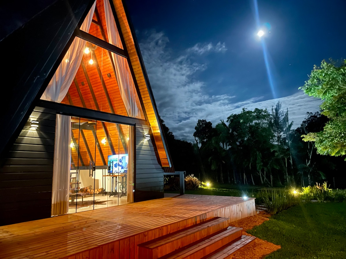The cabin exterior is illuminated at night, showcasing its A-frame design with high ceilings and large glass windows. A wooden deck leads to the entrance, surrounded by lush greenery. The moonlight adds a serene ambiance to the scene.