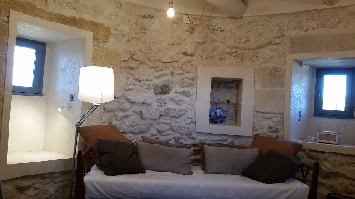 A cozy seating area features a simple daybed adorned with soft cushions. The stone walls provide a rustic charm, complemented by two small windows allowing natural light to enter. A standing lamp and wall light offer additional illumination, enhancing the inviting space.