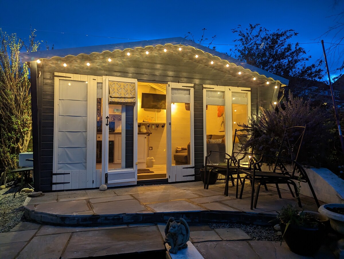A rustic cabin is illuminated by string lights at dusk, showcasing a welcoming entrance. The patio features several chairs arranged around a stone area. Inside, warm light highlights the kitchenette and living area, creating a cozy atmosphere for relaxation.