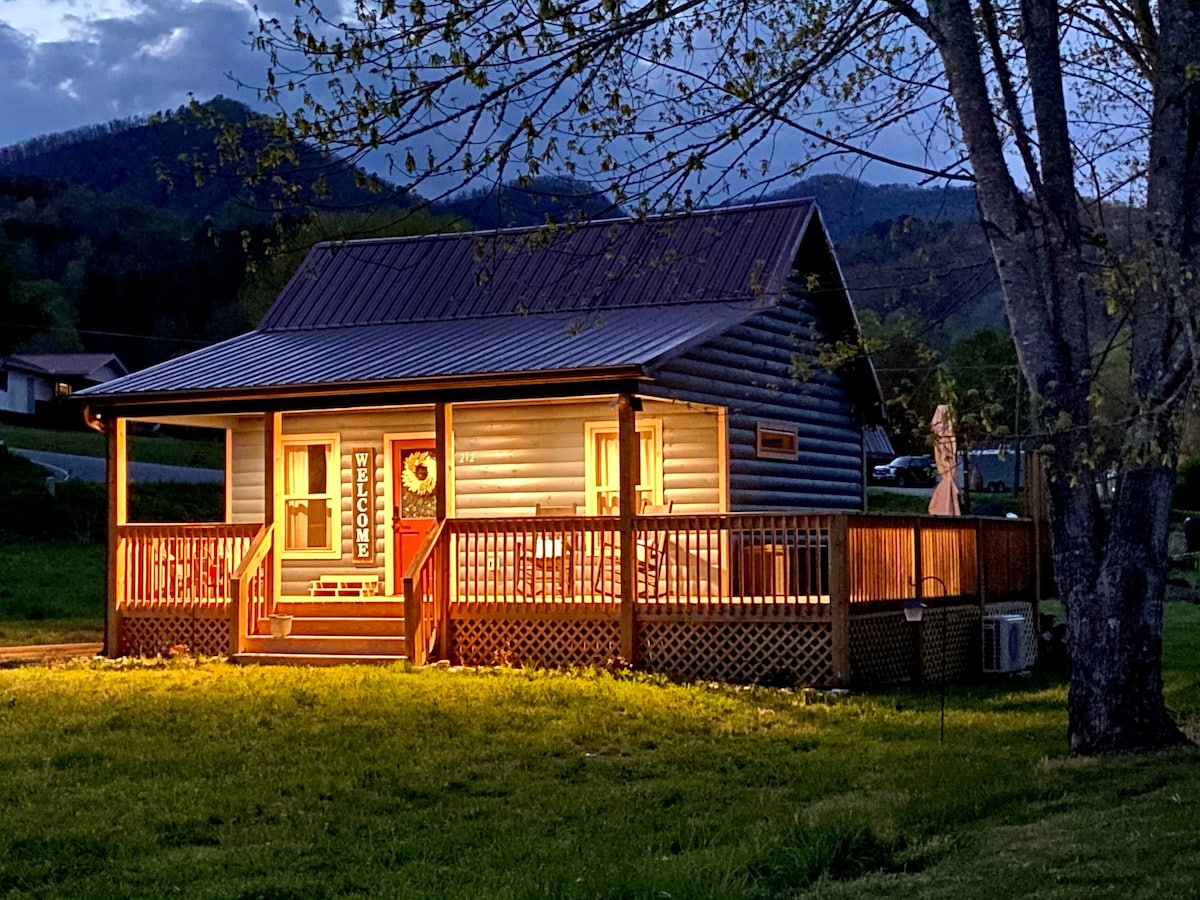 The cabin's exterior is softly illuminated during twilight, highlighting the inviting porch with a comfortable seating area. Surrounded by grassy terrain and framed by mountains, the rustic log structure features a pitched roof and large windows, creating an appealing presence in the landscape.