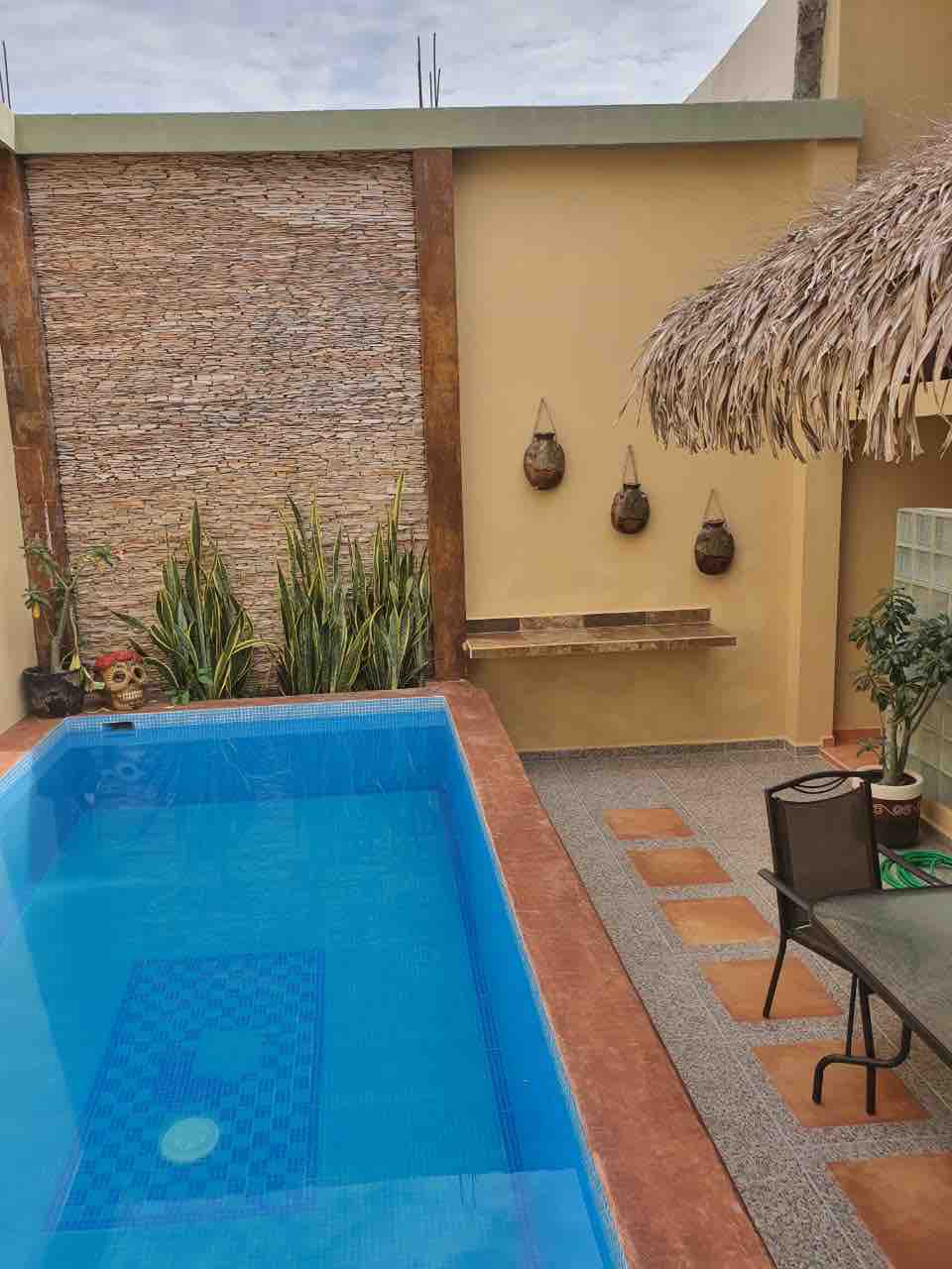 A sparkling private pool is bordered by a textured stone wall and tropical plants. A shaded seating area is positioned nearby, featuring a small table and chair. Decorative wall hangings add a touch of interest to the serene outdoor space, inviting relaxation.