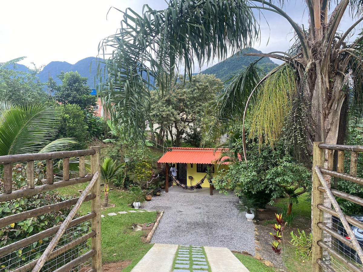A gravel pathway leads to a yellow house with a red roof, surrounded by lush greenery and tall palm trees. The backdrop features rolling mountains under a partly cloudy sky, creating a serene and natural atmosphere.