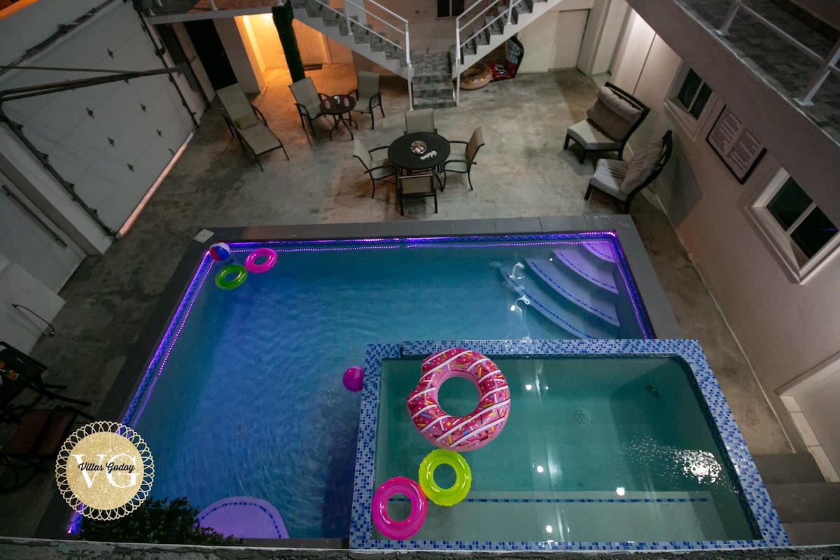 An aerial view of a shared pool area featuring a main pool with colorful inflatable floats and a connected jacuzzi. Surrounding seating is arranged, including a table with chairs, set against a backdrop of modern architecture.