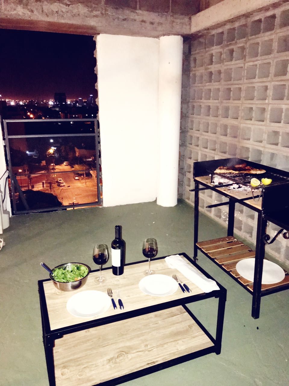 A private terrace space is presented, featuring a charcoal grill alongside a small dining table set for two. Two glasses of red wine sit next to a salad bowl on the table. The backdrop showcases city lights visible through a large window.