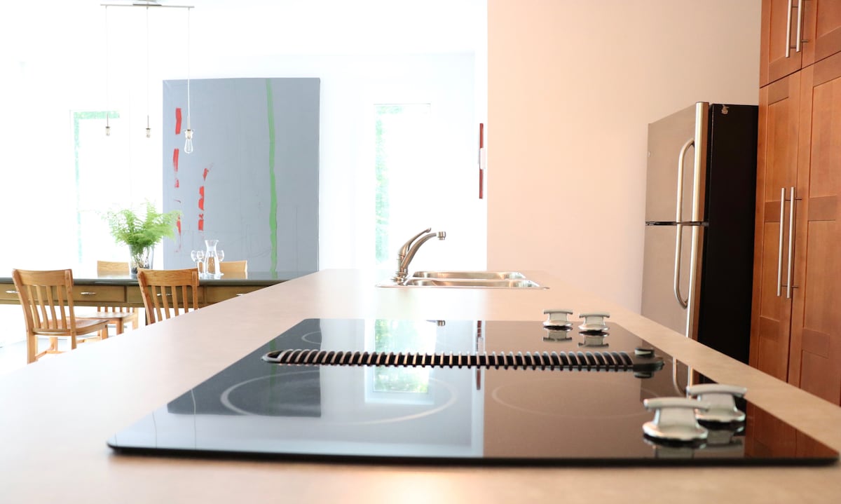 A modern kitchen is showcased, featuring a sleek cooktop integrated into a spacious countertop. Behind, wooden cabinetry complements a stainless steel refrigerator. A dining area with a wooden table and chairs is visible in the background, illuminated by natural light through large windows.