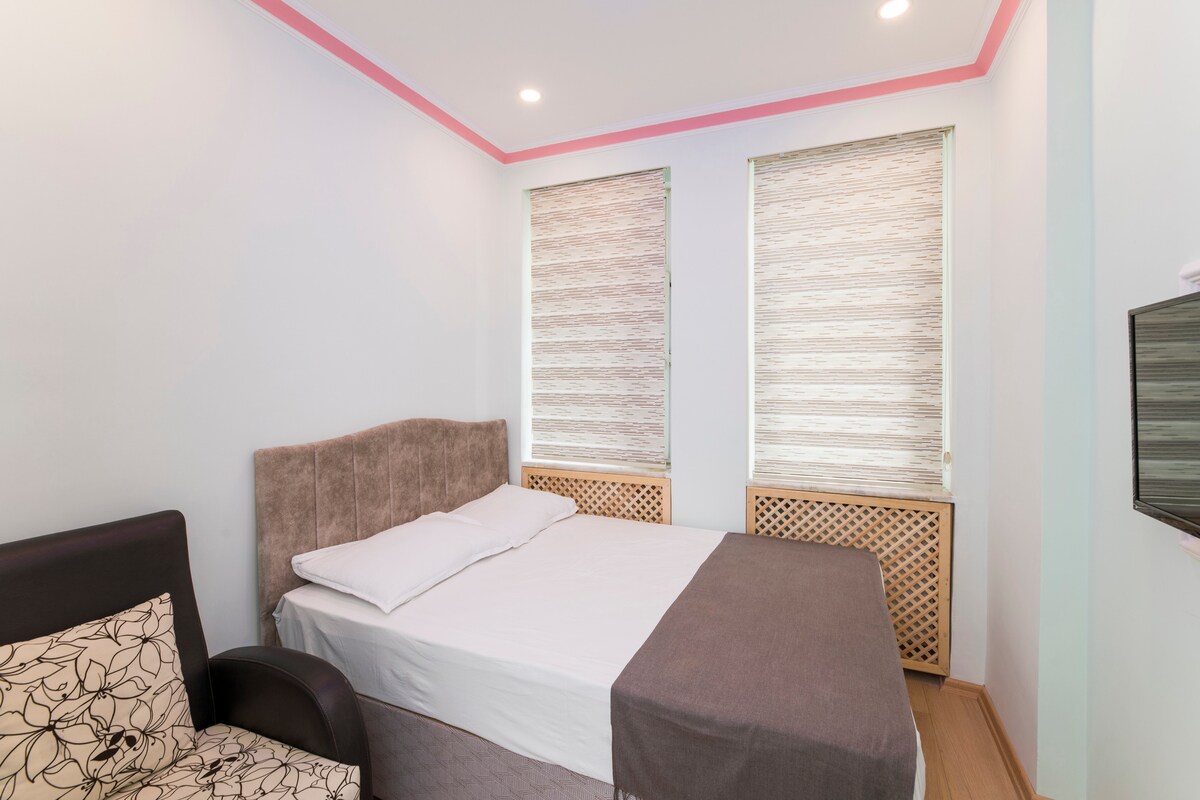 A comfortable bedroom features a spacious bed adorned with crisp linens. Natural light enters through two stylish window shades, illuminating the room. A cozy armchair provides a seating option, complemented by a modern wall-mounted television.