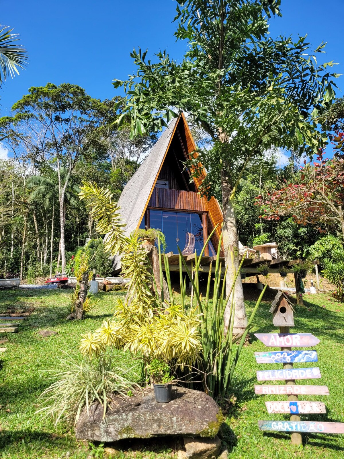 An A-frame cabin is situated amidst lush greenery, featuring large glass windows that invite natural light. The exterior displays a mix of wood and modern elements, with various plants enhancing the surrounding landscape. A signpost nearby offers welcoming messages.