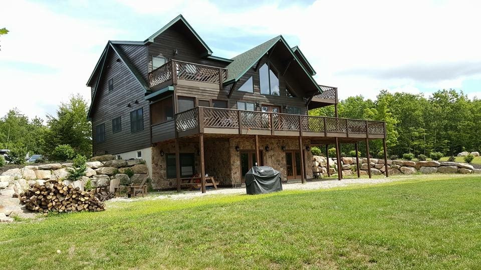 The mountain retreat is set against a backdrop of lush greenery, featuring a spacious two-story structure with a combination of wood and stone exteriors. Multiple decks with railings provide access to outdoor seating. A grill stands on the patio, complemented by stacked firewood.