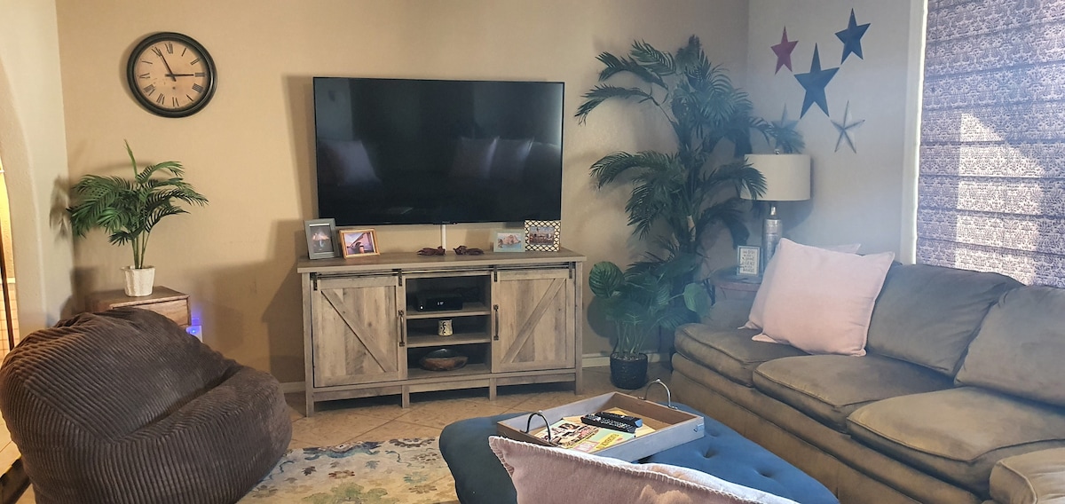The living room features a spacious sectional couch and a large flat-screen TV mounted above a media console. A cozy bean bag chair is positioned nearby. Natural light filters through patterned blinds, illuminating decorative plants and a wall adorned with colorful stars.