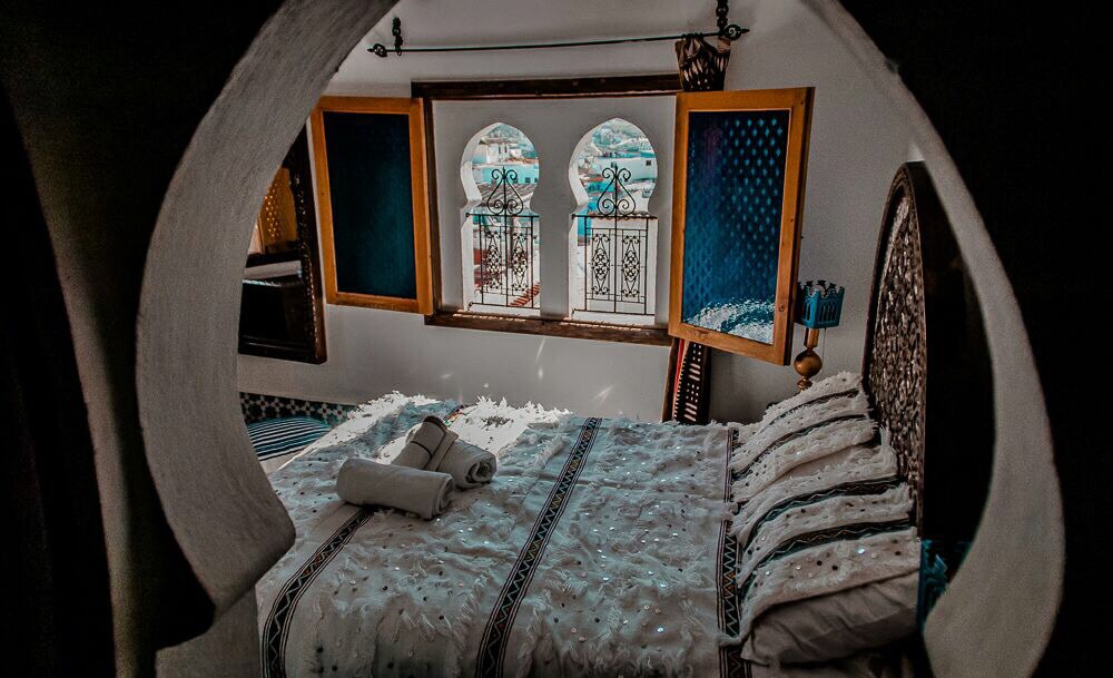 A cozy bedroom features a spacious bed adorned with a decorative white blanket and several patterned pillows. The windows, framed in arched alcoves, showcase scenic views of the outside. Natural light enters through the yellow shutters, creating a calm environment.