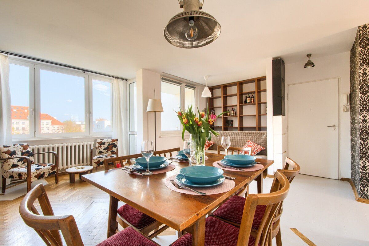 A dining area features a wooden table set with blue plates and wine glasses, surrounded by chairs. Natural light floods in through large windows, illuminating the space. A cozy seating area with patterned armchairs is visible in the background, creating an inviting environment.