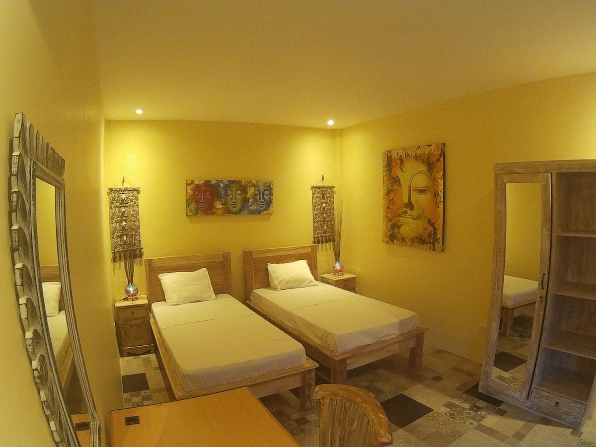 A warm room is presented with two twin beds positioned beside each other. Soft yellow walls enhance the space, complemented by artistic wall decor. A large mirror reflects the room's interior, while a wooden wardrobe and a desk add functional elements, offering a cozy and comfortable environment.