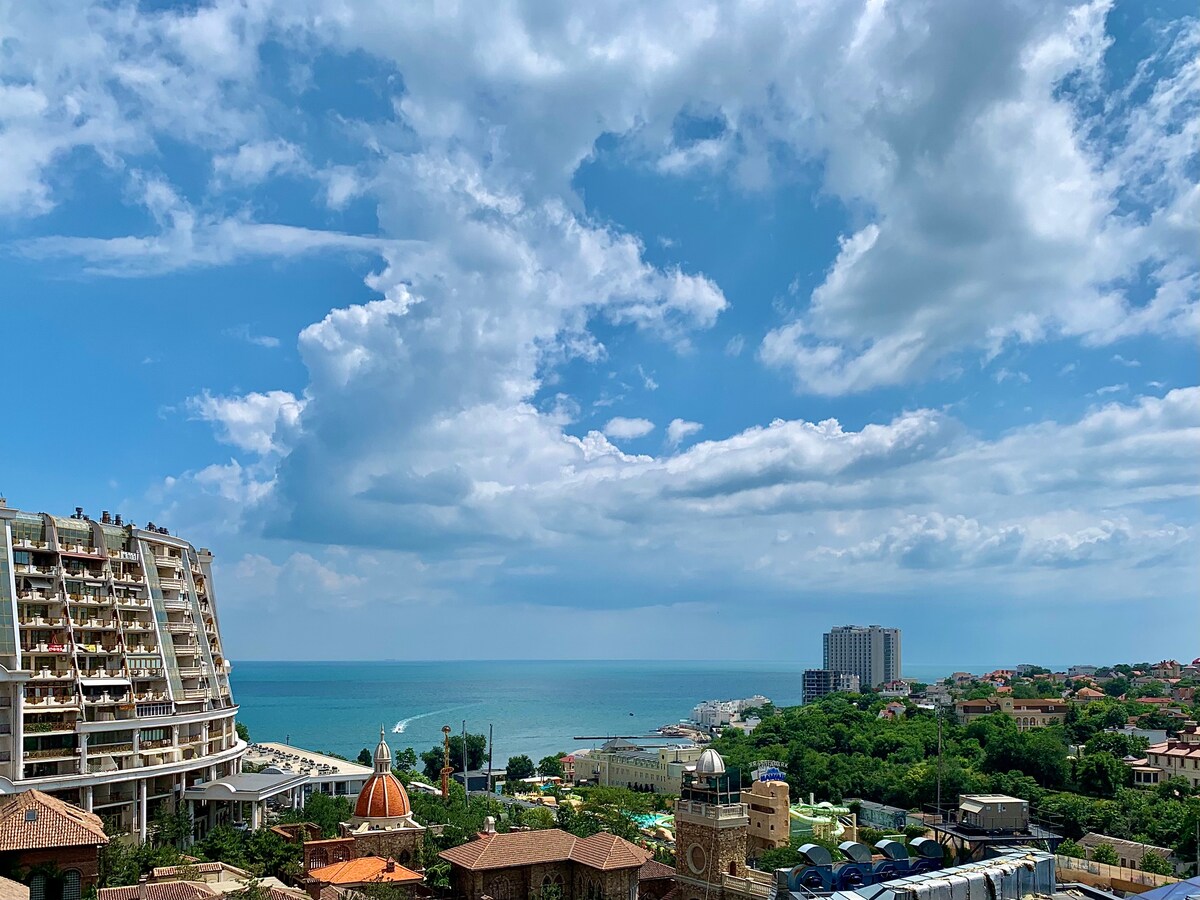 A panoramic view captures a serene coastline with soft waves lapping against the shore. The skyline features a mix of modern buildings and traditional architecture, while fluffy clouds drift across a bright blue sky.