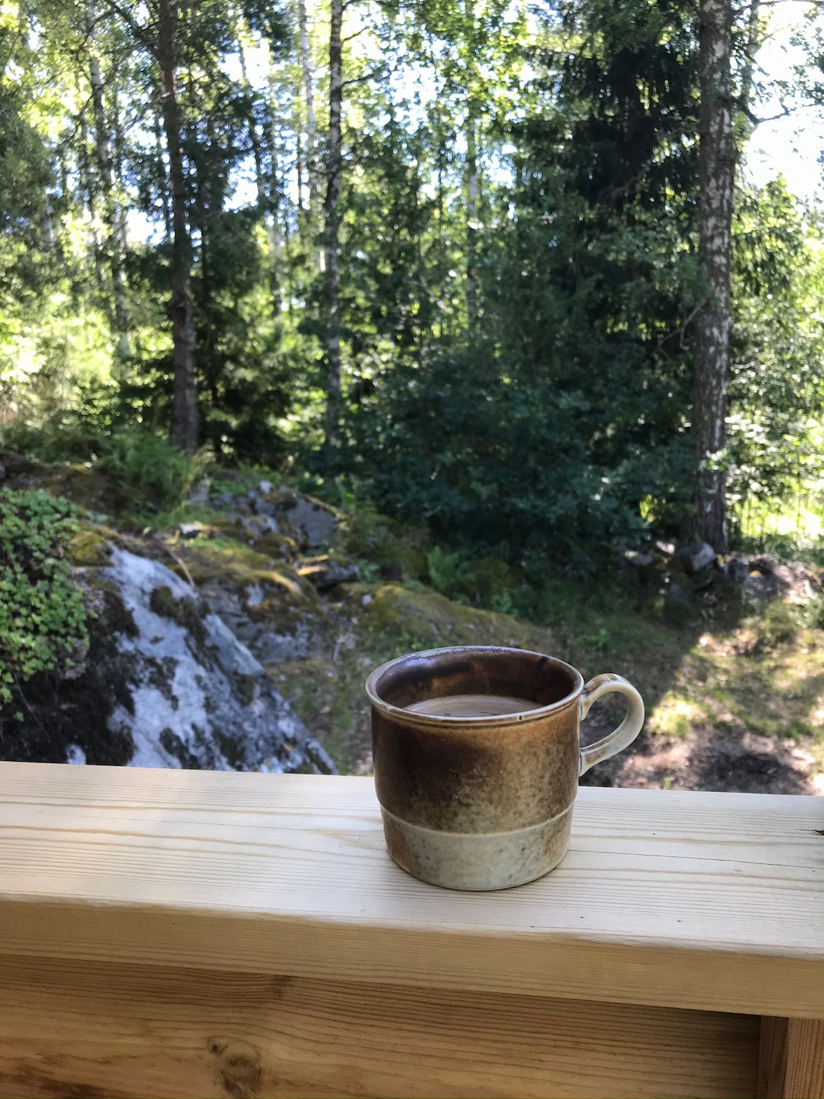 A rustic ceramic mug filled with coffee is placed on a wooden ledge. The background features a lush green forest, with trees and rocky terrain visible, creating a serene outdoor ambiance.