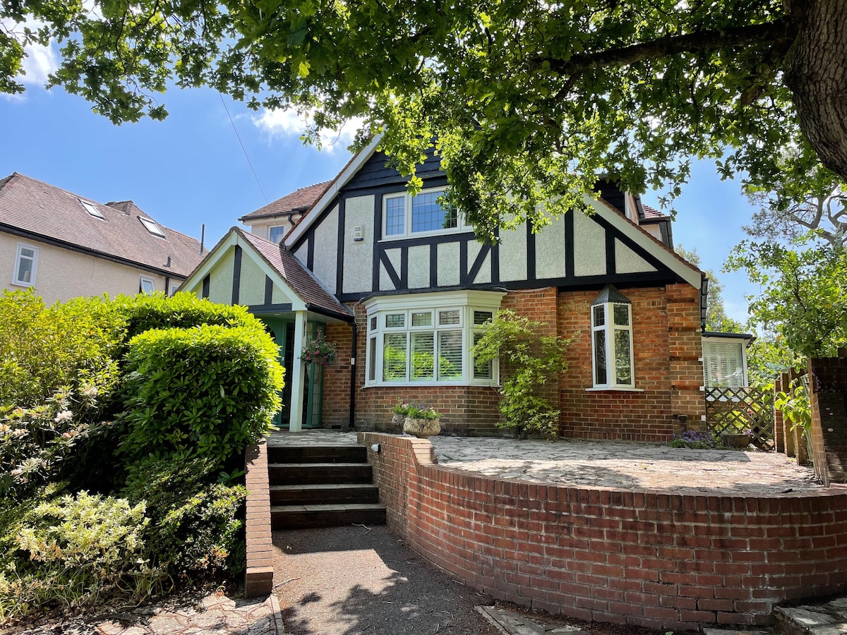 The exterior of a charming house is showcased, featuring a combination of traditional timber framing and brickwork. A well-manicured garden surrounds the entrance, with leafy green trees providing shade. Steps lead to a welcoming doorway, accentuated by a decorative entrance awning.